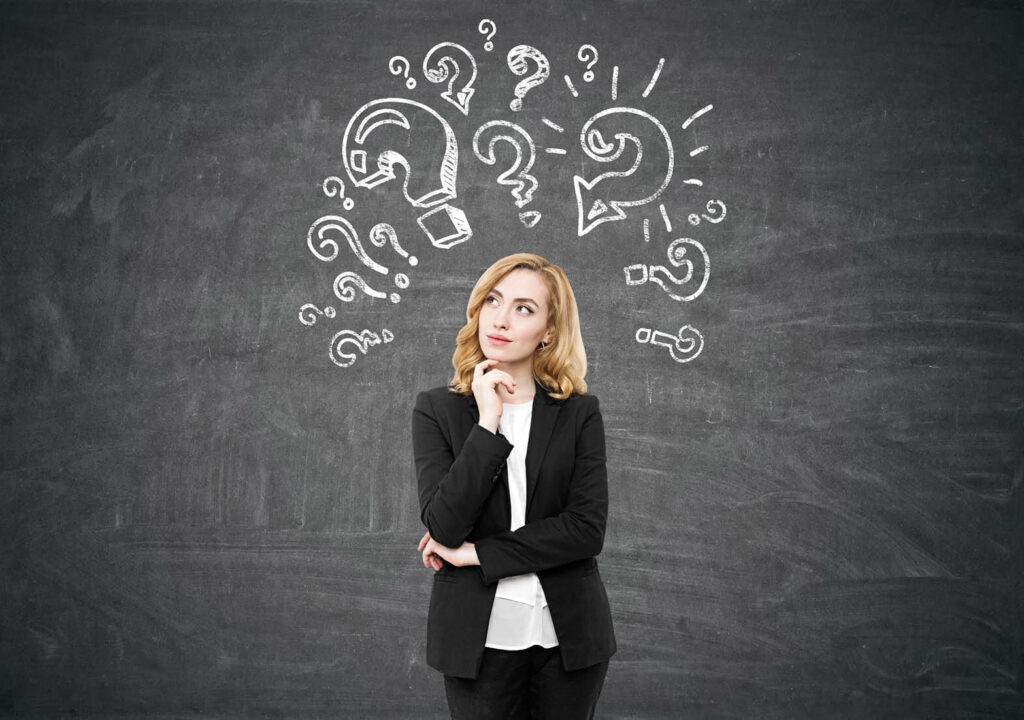 Pensive businesswoman with red hair is standing near a chalkboard with question mark sketches on it. Concept of looking for the answer. Mock up