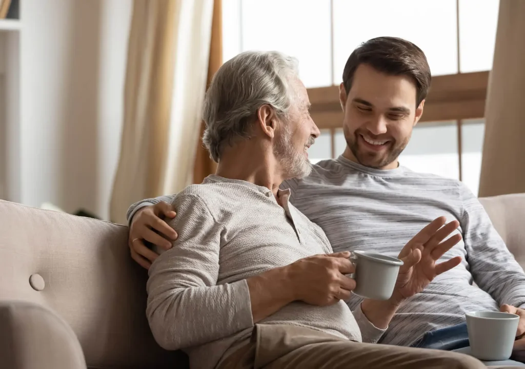 Joyful two generations family sharing news with cup of tea. Happy middle aged grey haired men relaxing on cozy sofa with smiling grownup son, spending leisure weekend morning time with cup of coffee tea. Joyful two generations family talking sharing news.