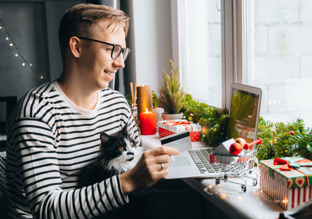 happy young man with funny cat holding credit debt card shopping online purchase xmas presents on laptop home. Christmas gift box, New Year winter sale discount. Safe ordering delivery at quarantine
