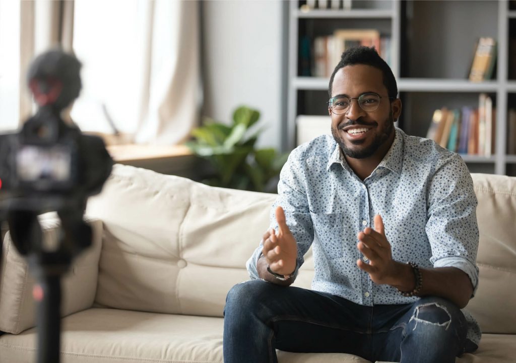 Happy young african american man speaker in eyewear sitting in front of camera, recording educational video webinar training at home. Smiling skilled biracial guy introducing for job interview.
