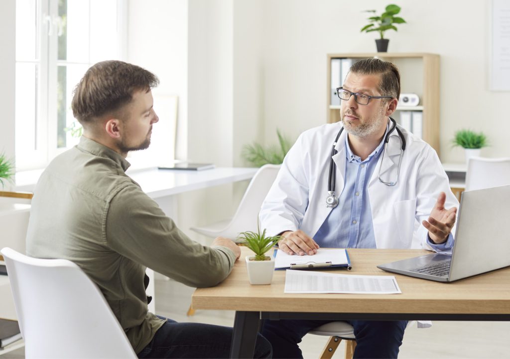 Male general practitioner clinic doctor consulting mature patient at appointment, explaining, showing medical checkup results on laptop screen, serious physician giving recommendations at meeting
