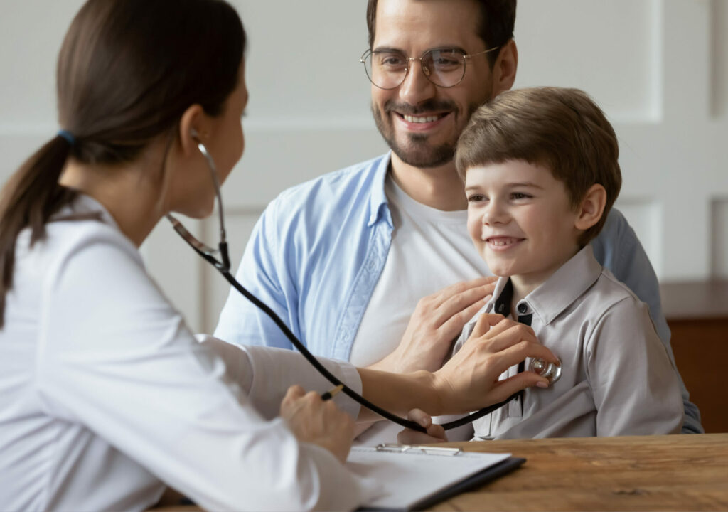 Female pediatrician listen to smiling 6s boy patient heart chest with stethoscope during visit with dad to clinic. Woman doctor examine small child at consultation with father. Healthcare concept.