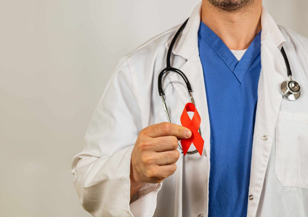 Medical professional holds a red ribbon symbolizing HIV awareness warmly. A healthcare professional with a stethoscope wears a white coat and blue scrubs, holding a red ribbon that represents HIV awareness and support during a health awareness campaign.
