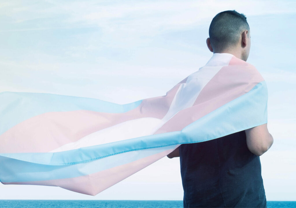 closeup of a person, seen from behind, wrapped in a transgender pride flag outdoors, with the blue sky in the background, in a panoramic format to use as web banner or header