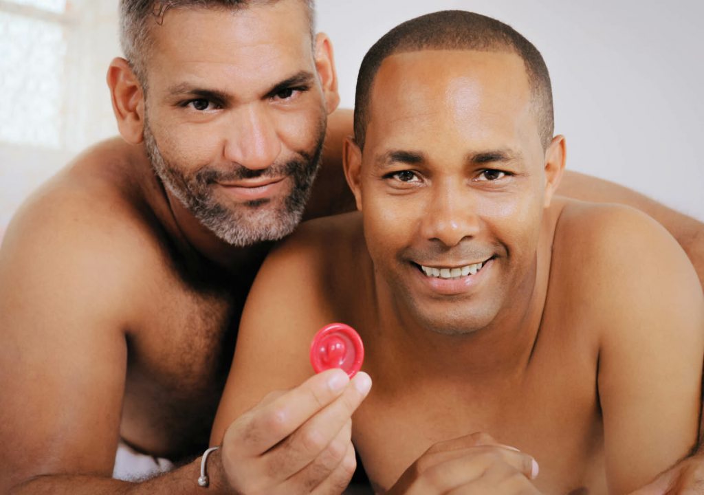 Homosexual couple, happy gay people smiling at camera. Hispanic friends in bedroom showing condom for safe sex.