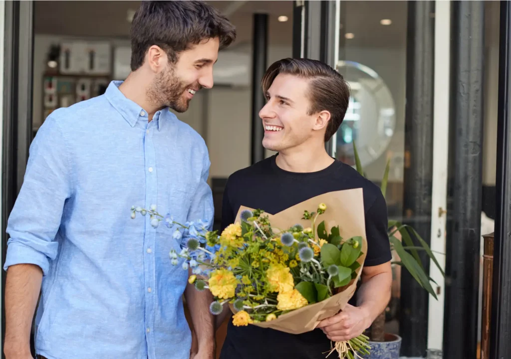 Loving Male Gay Couple Holding Hands Coming Out Of Florists Holding Bunch Of Flowers