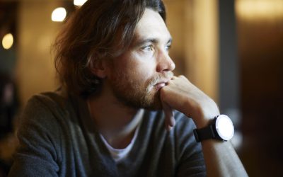 Portrait of handsome hipster guy with long hair and beard sitting in sunny cafe, resting near window. Romantic man looks lonely.