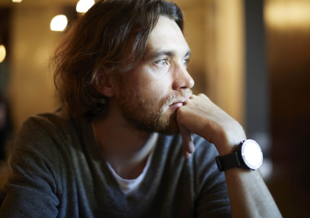 Portrait of handsome hipster guy with long hair and beard sitting in sunny cafe, resting near window. Romantic man looks lonely.