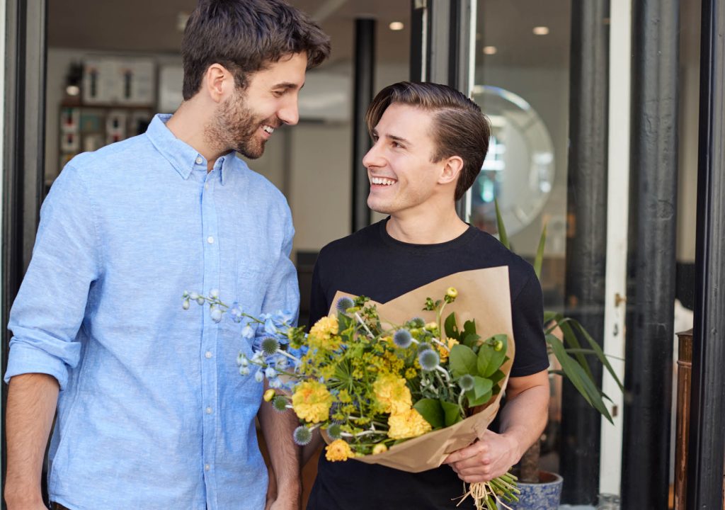 Loving Male Gay Couple Holding Hands Coming Out Of Florists Holding Bunch Of Flowers