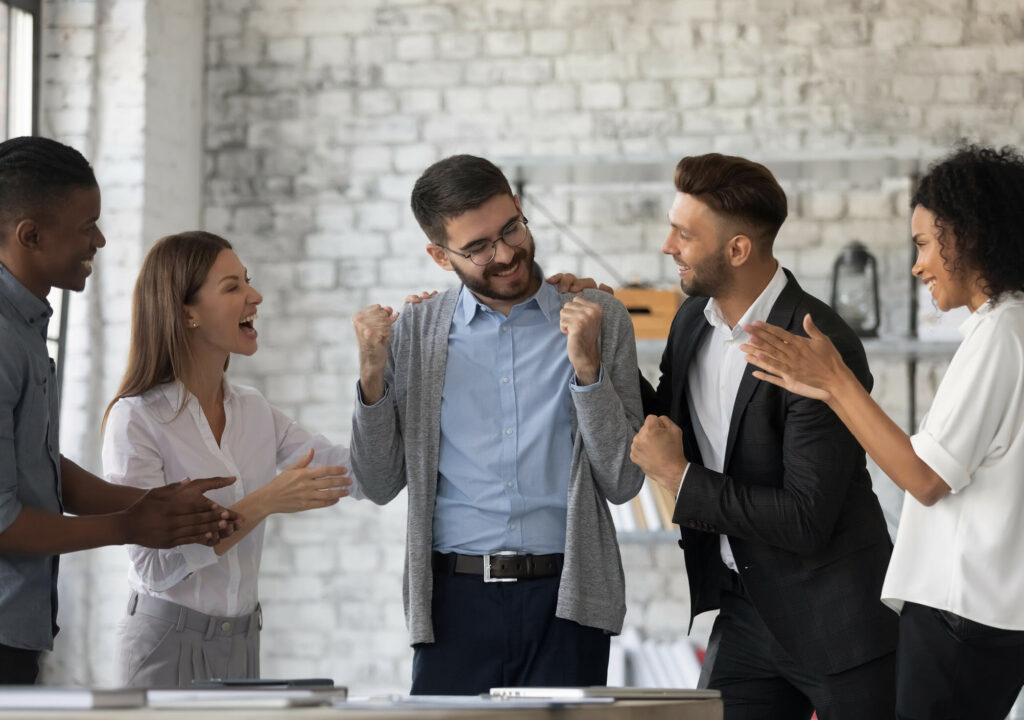 Smiling multiracial businesspeople congratulate colleague with job success or achievement. Happy supportive diverse multiethnic employees greet excited male worker with work promotion.