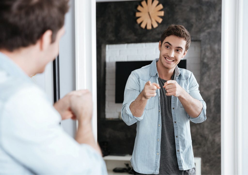 Smiling young man pointing fingers at his mirror reflection while standing at home