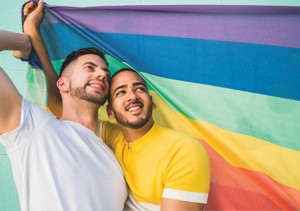 Portrait of young gay couple embracing and showing their love with rainbow flag at the street. LGBT and love concept.