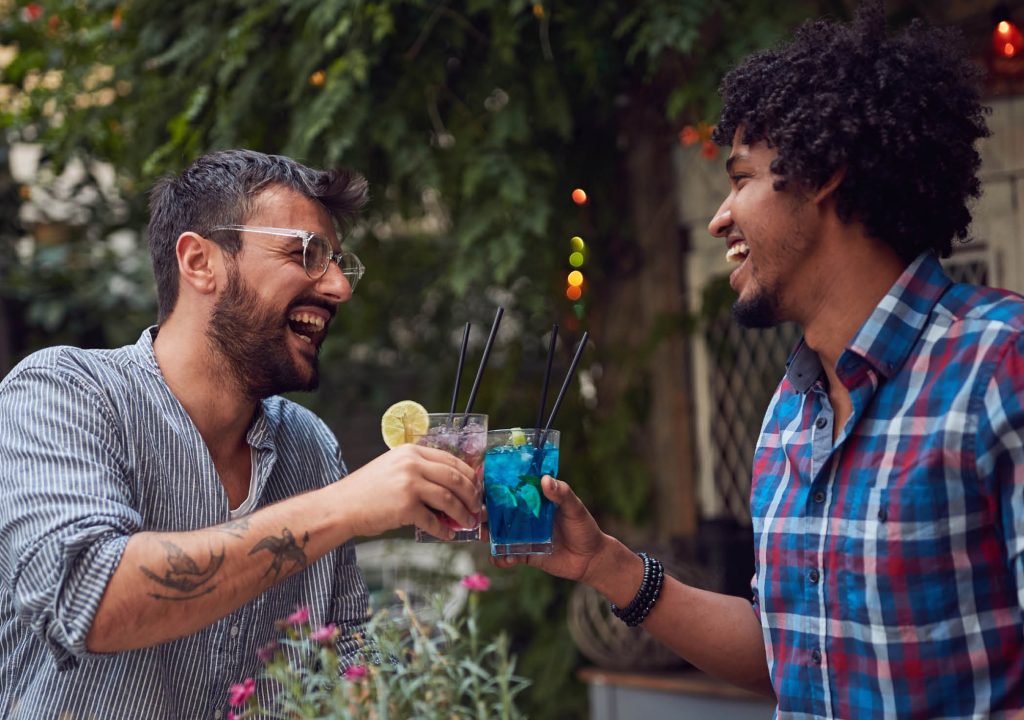 Two young multiethnic male laughing and toast with cocktails in outdoor cafe. multiethnic, friendship, socializing, leisure, casual