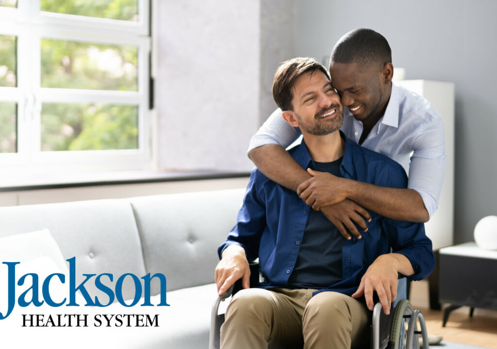A male patient is In Wheel Chair with his partner beside him. Jackson health logo on the bottom left of the photo