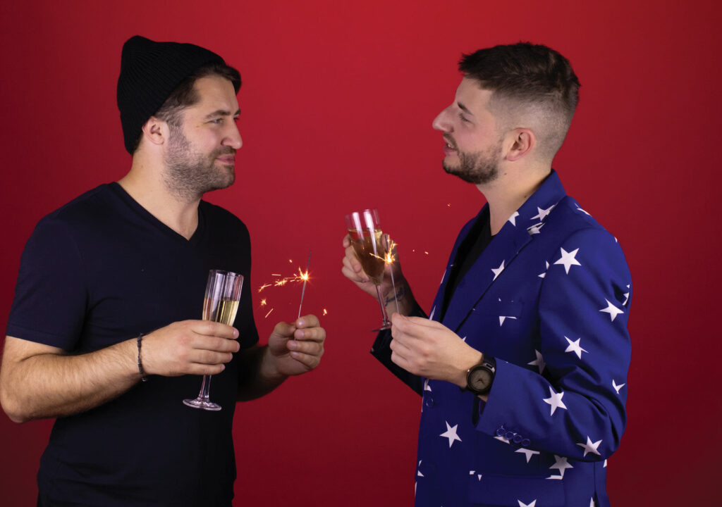 Two attractive stylish gay men celebrating New Year party, homosexual couple holding sparklers and glasses with champagne congratulating each other on red background. Romantic Christmas concept