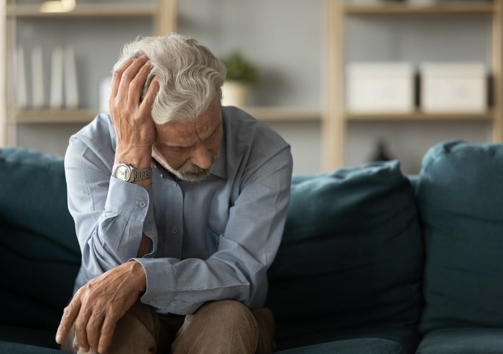 Frustrated unhappy middle aged mature man sitting on sofa, feeling depressed alone at home. Confused senior retired grandfather worrying about difficult life decision, copy space, old people solitude.